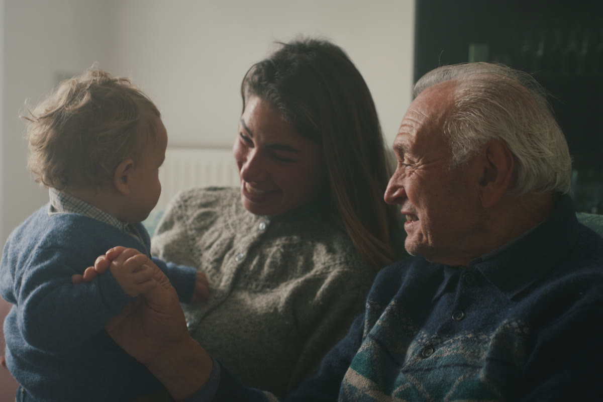A grandfather, his daughter and her baby son sitting together on a sofa at home.