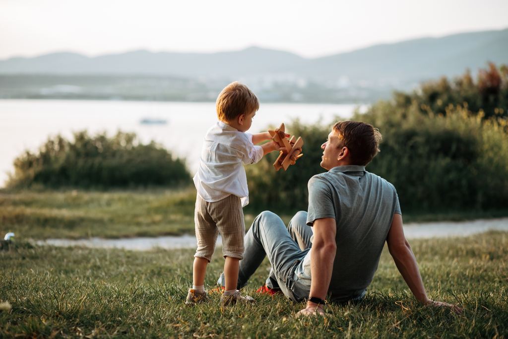 A father sitting on the grass beside a lake, watching his young son hold up a wooden toy aeroplane.