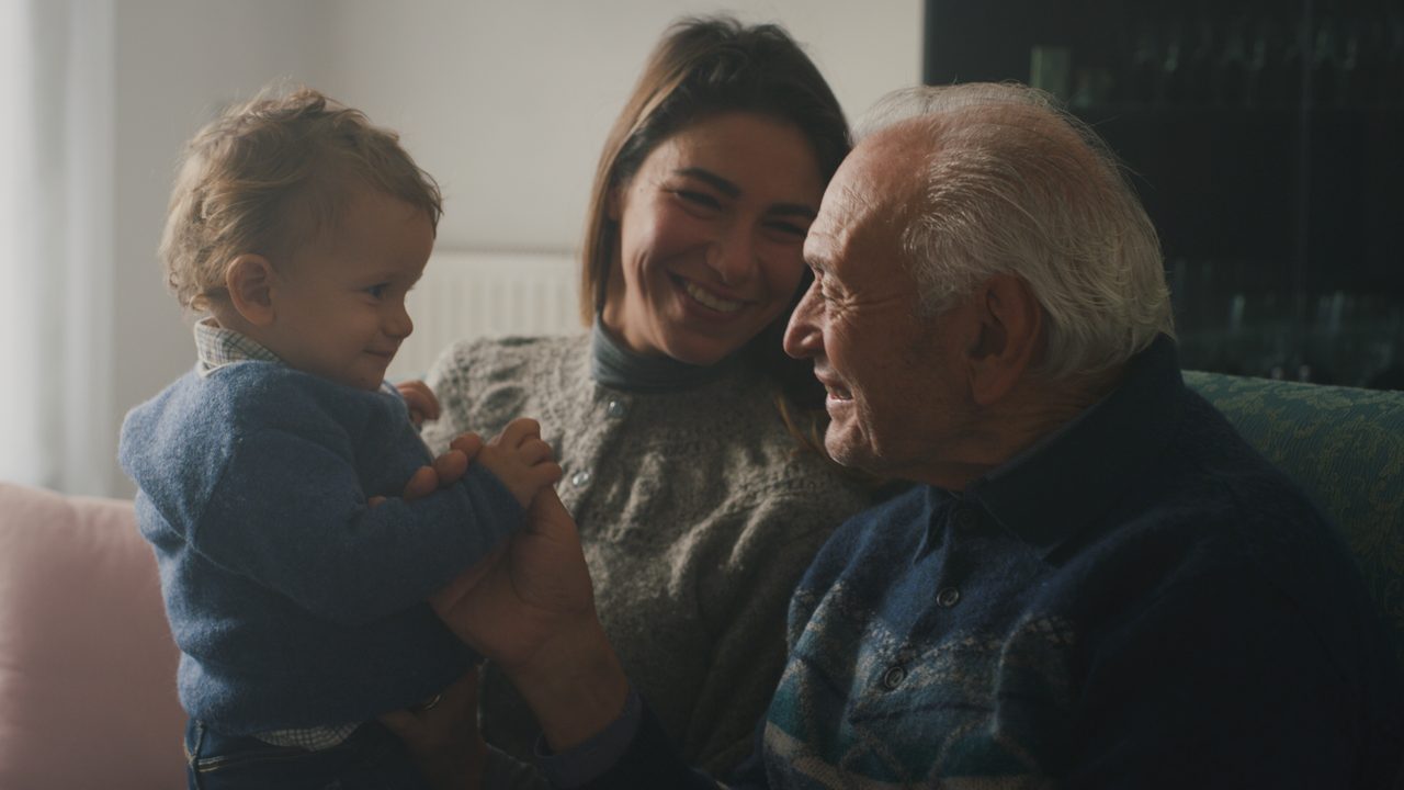 A grandfather, his daughter and her baby son sitting together on a sofa at home.